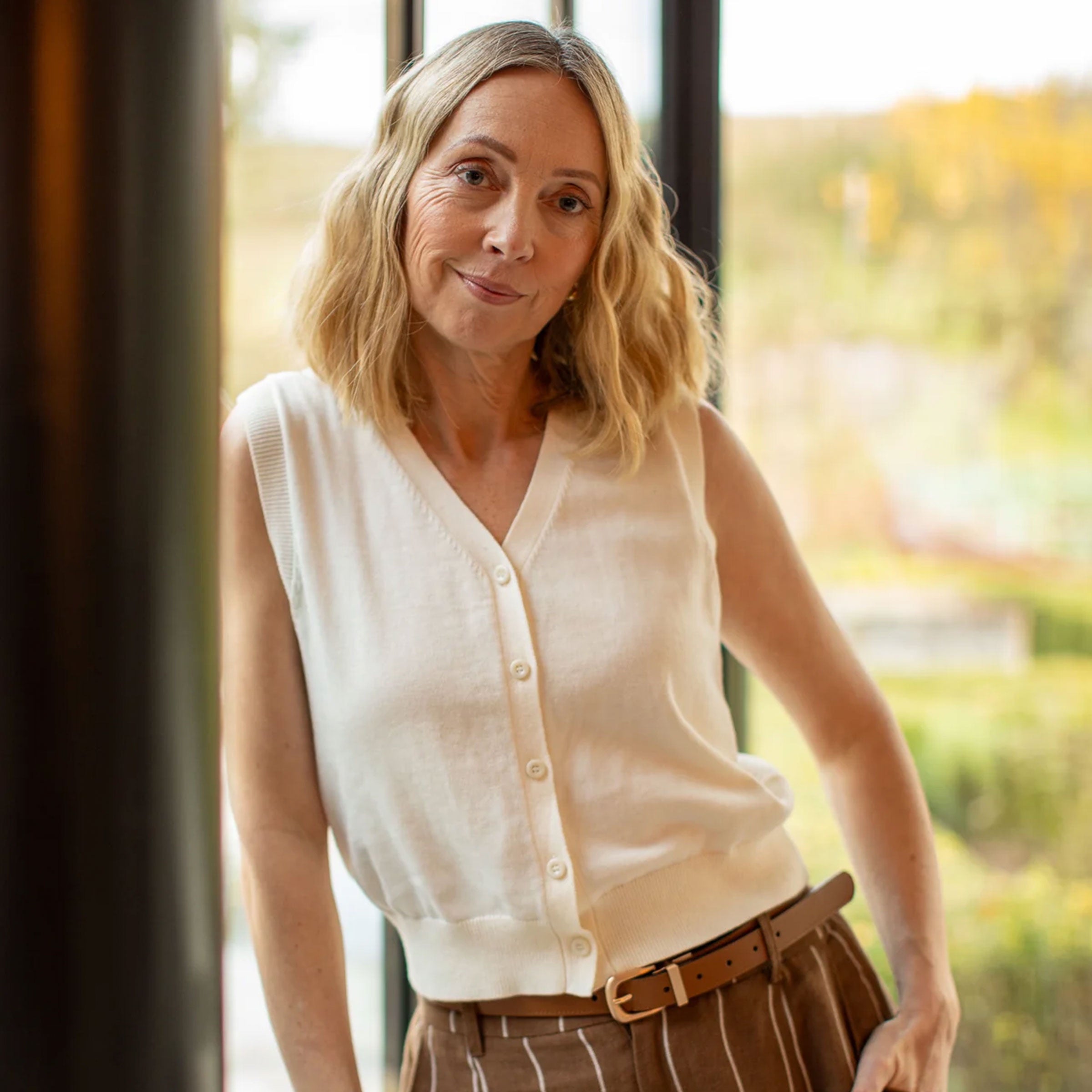 Woman wearing a white sleeveless blouse and brown pants standing indoors with a blurred outdoor background.